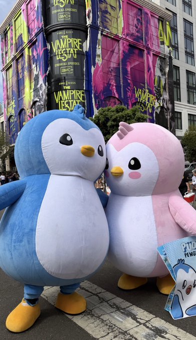 Two large inflatable mascot costumes, a blue penguin and a pink penguin, stand on a city street with a billboard for 'THE VAMPIRE FIESTA' and 'AMC Original Episodes' visible in the background.