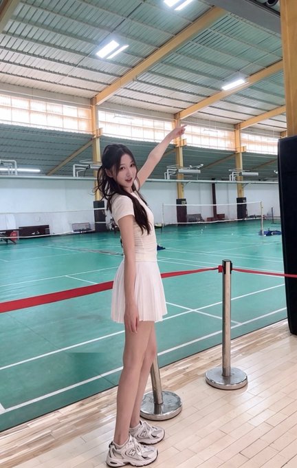 A young woman with long dark hair posing in a white pleated skirt and white top on what appears to be a badminton court, pointing upward with one arm, wearing chunky white sneakers.