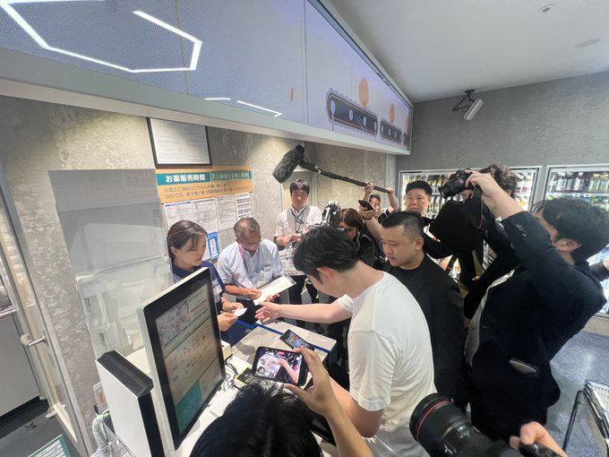 A crowd of press and onlookers, including photographers with boom microphones, gathers around a LAWSON convenience store counter where staff assist customers with the first-ever JPYC cryptocurrency payment trial in Japan. A sign reading "印象版画特価" with the time "11:00-19:00" is visible on the counter as attendees document the historic transaction.