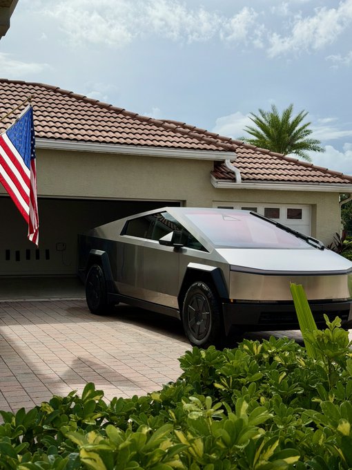 A silver Tesla Cybertruck parked in a driveway in front of a house with a tile roof, with an American flag displayed and palm trees in the background.