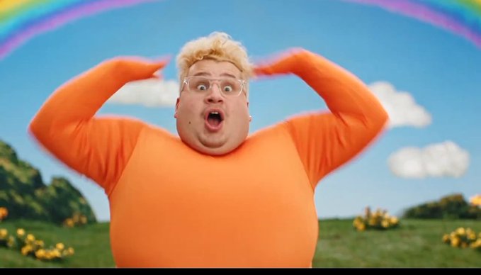 A man with blonde hair and glasses, wearing an orange shirt, stands outdoors with a shocked expression, his hands raised near his head, against a backdrop of a blue sky with clouds and a rainbow.