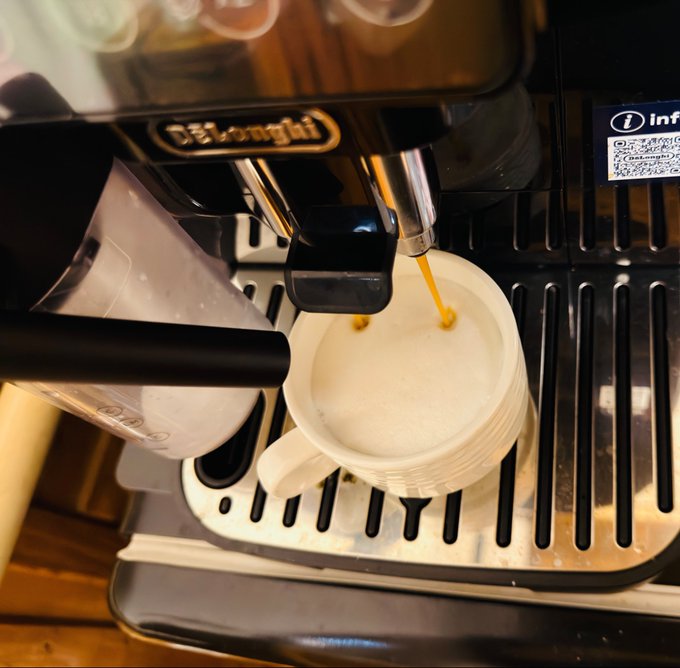 The image shows a coffee machine pouring milk into a white cup placed on top of a metal tray. The scene takes place on a wooden table, which adds warmth and coziness to the setting. The focus of the image seems to be on the process of making coffee or tea using this particular coffee maker.