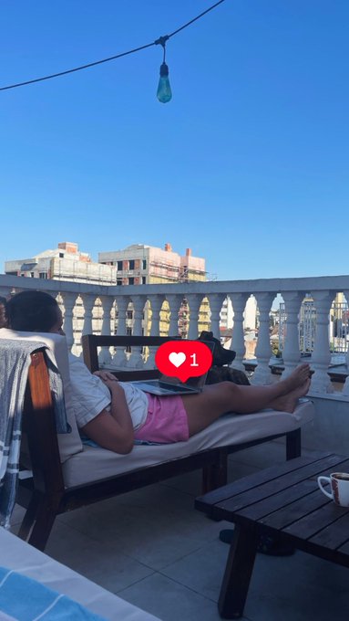 A man relaxing on a rooftop lounge chair with a laptop, showing a '1' heart reaction notification, with a cityscape and clear blue sky in the background.