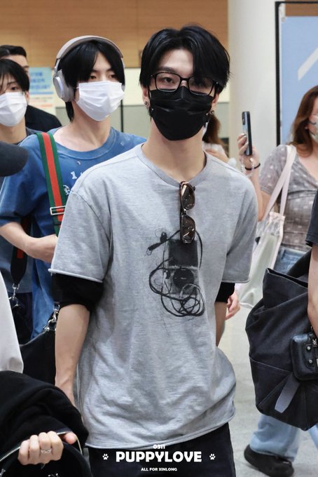 A person wearing a black mask, glasses, and a gray graphic T-shirt with a black mask hanging from it stands at what appears to be an airport, accompanied by others wearing masks and headphones. The watermark "PUPPYLOVE ALL FOR XINLONG" at the bottom of the image ties the photo to the fan account's hashtags referencing #XINLONG (씬롱/シンロン/鑫隆) and #ALPHADRIVEONE, suggesting this is an airport arrival photo of the K-pop idol.