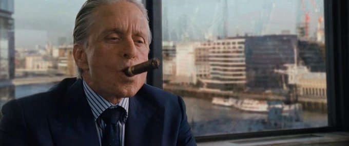 A still of an older man in a suit smoking a cigar, looking out a window at a harbor with construction cranes.