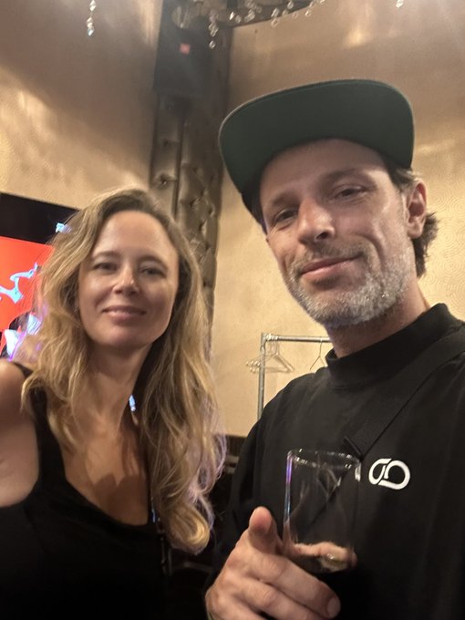 A selfie of a woman with long blonde hair in a black top and a man wearing a dark cap and black shirt holding a glass.