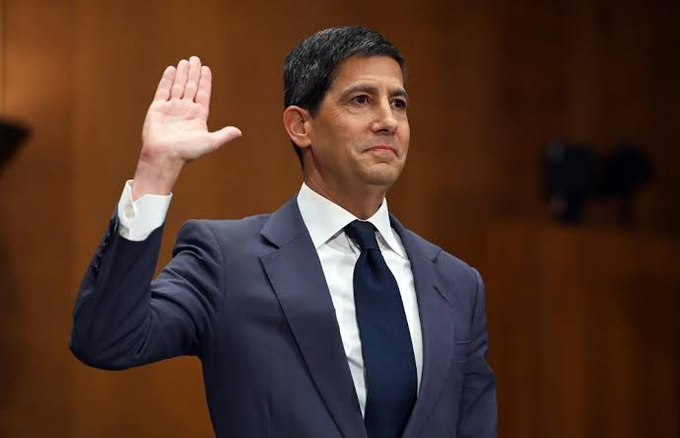 A man in a dark blue suit raises his right hand as if being sworn in, in what appears to be a Senate hearing room.