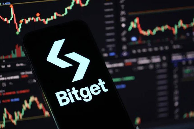 Close-up of a smartphone displaying the Bitget cryptocurrency exchange logo, positioned in front of a computer screen showing a candlestick trading chart with red and green price movements.