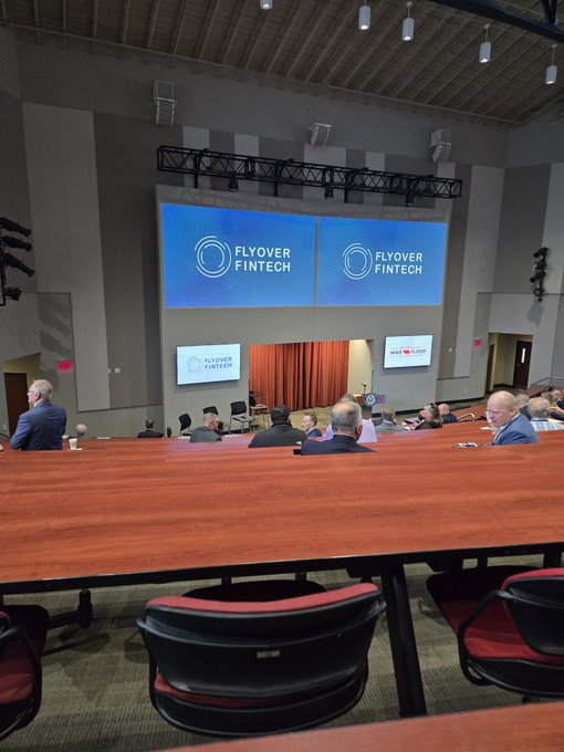 A view from the back of a conference room where attendees are seated at tables facing a stage with large screens displaying "Flyover Fintech" logos. The setting suggests a fintech industry event, aligning with the post's reference to Telcoin ($TEL) in attendance.