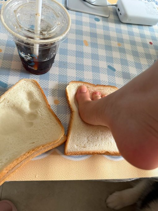 A foot rests playfully between two slices of bread on a blue-checkered tablecloth, next to a soda with a straw, captioned "toestie with my freddo."