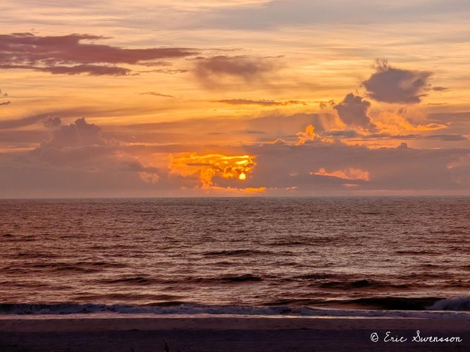 A serene sunset over the ocean with golden and orange hues reflecting on the waves, signed '© Eric Swenson' in the bottom right corner.