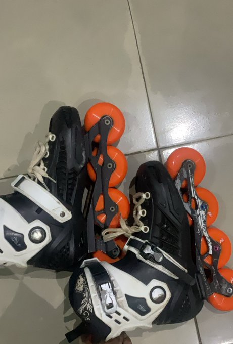 A pair of black and white inline skates with orange wheels sit on a tiled floor, showing some wear and scuffs.