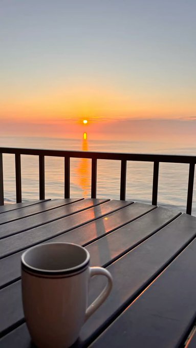A peaceful sunrise scene over a calm ocean viewed from a balcony, with a white coffee mug sitting on a wooden table in the foreground as the sun's reflection shimmers on the water.
