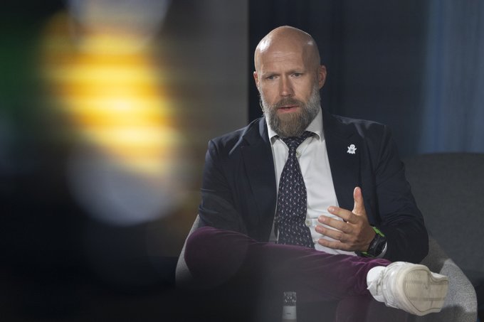 A bald, bearded man in a dark suit and patterned tie sits in a chair gesturing with his hand while speaking, appearing to be engaged in a panel discussion at a formal conference setting. He matches the description of CEO F. Gregaard participating in a panel at the Hamburg Sustainability Conference alongside UNDP, EY, and Stellar representatives about using public blockchain infrastructure for development funding.