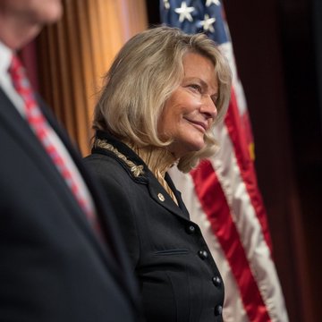 Senator Cynthia Lummis stands beside an American flag, smiling in what appears to be an official government setting, aligning with her public push for the Senate to pass the CLARITY Act on crypto market structure.