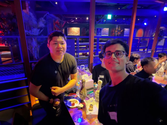 Two men pose for a selfie at a dimly lit bar with blue lighting; one wears glasses and a black shirt while the other wears a dark t-shirt.
