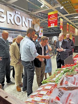 The image shows a group of people standing around a table filled with food items inside a grocery store. The customers are looking at the products displayed on the table, possibly deciding which items they want to purchase. There are several bags of chips visible among other food items available for sale. The scene captures a moment in time where shoppers are browsing and making choices about their purchases.