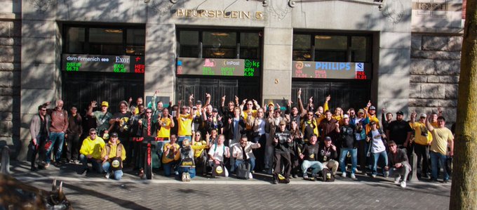 In this image, there is a large group of people standing next to each other outside an office building. The individuals are wearing different types of clothing, but they all seem to be participating in something together. There's also some text visible on the photo, which appears to discuss investing in Bitcoin and shares insights about its potential as a viable long-term investment. This group of people could potentially represent a community or organization that is focused on cryptocurrency, or it may simply be an informal gathering outside the office building for various reasons related to crypto.