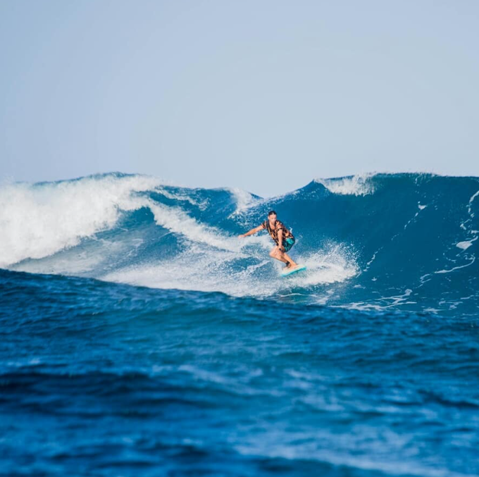 A surfer rides a large blue wave with white foam under a clear sky.