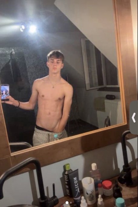 Mirror selfie of a young shirtless man in grey sweatpants standing in a bathroom, holding a phone; various skincare and toiletry bottles are visible on the counter.