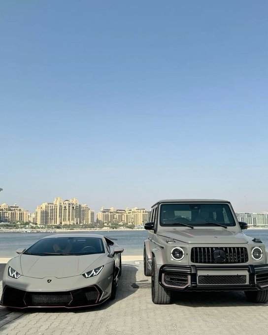 The image features two luxury cars parked next to each other, with a cityscape in the background. The first car is a silver sports car, while the second car is a black SUV. Both vehicles are positioned facing the same direction, as if ready for their owners to embark on an adventure or attend a special event. The cityscape behind them adds an urban touch to the scene, indicating that this location could be in Dubai or another city with a bustling transportation network and modern architecture.