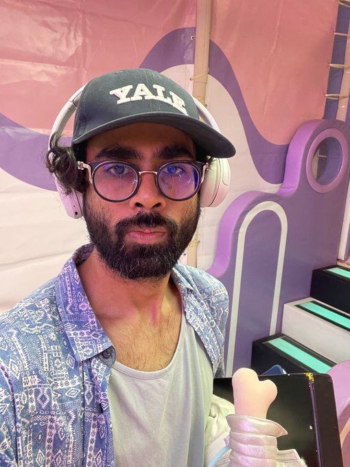 A bearded man wearing a green "YALE" cap, purple-tinted glasses, and white headphones poses in front of a pink and purple backdrop with abstract designs. The caption reflects the post's theme of exploring diverse creative pursuits, capturing a moment that suggests versatility and the search for a fitting creative identity.