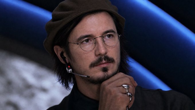 A man with shoulder-length dark hair, a mustache, and goatee wears a brown knit beanie and thin wire-rimmed glasses while resting his chin on a ring-adorned hand, with a headset microphone near his mouth. The contemplative pose against a dark backdrop with blue lighting visually evokes the calculated, scheming tone of planning manipulative market tactics.