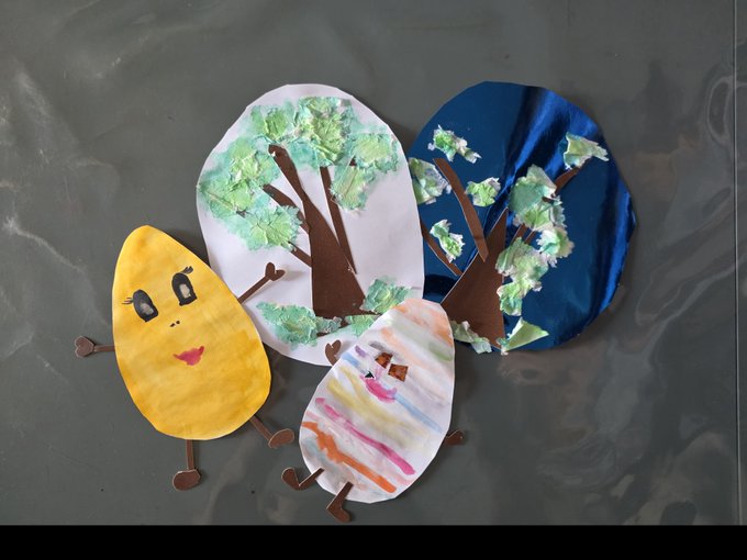 A child's craft project on gray paper showing paper plate trees with crumpled tissue paper leaves and two smiling characters made from paper plates in yellow and rainbow stripes.