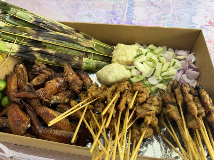 A spread of local Singaporean delights in a takeout box, featuring satay skewers, otah (spiced fish paste wrapped in banana leaves), and BBQ chicken wings with sambal and fresh cucumber-onion relish, perfect for a Token2049 private event catered by Hyperliquid.