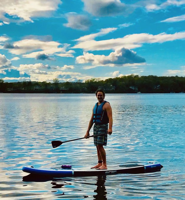 A man wearing a blue life vest stands on a stand-up paddleboard on a calm lake under a partly cloudy sky, holding a paddle.
