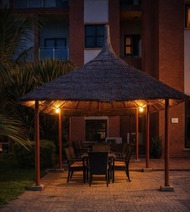 An illuminated thatched-roof gazebo houses a patio dining set with chairs and a table, situated in a courtyard surrounded by brick buildings and tropical plants at dusk.