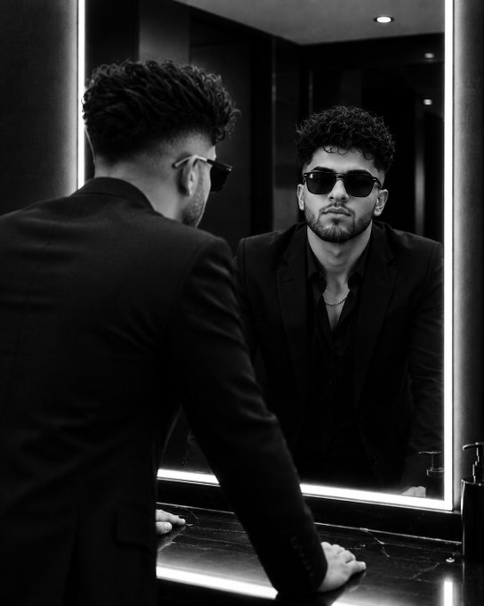 A black and white photograph captures a stylish man in a suit and sunglasses looking at his reflection in a backlit mirror.