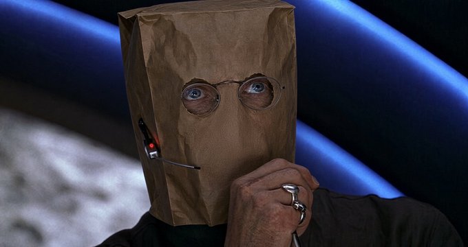 A person with a brown paper bag over their head, featuring cut-out eyes and drawn-on glasses, sits with a cigar protruding from the bag's mouth area, wearing a black shirt and silver rings.