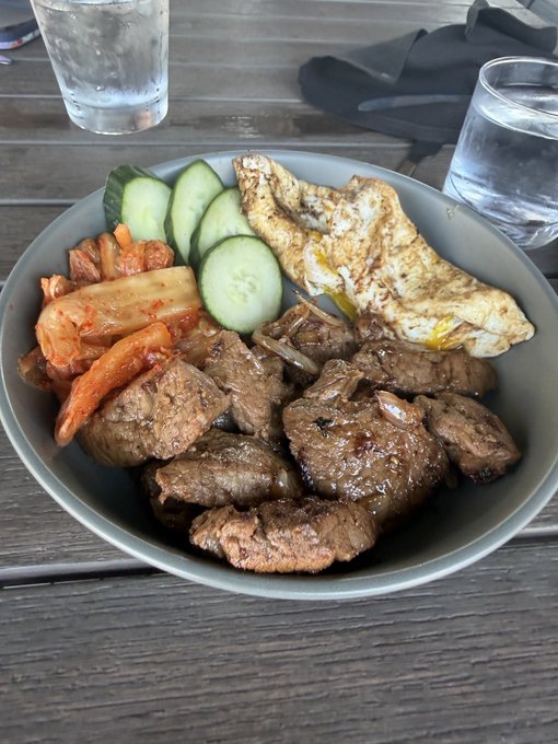 A gray bowl holds a meal of sliced beef, fried egg, cucumber slices, and kimchi, served alongside a glass of water on a wooden table.