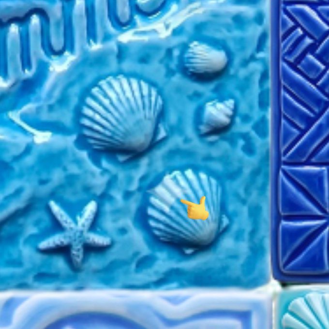 A close-up of a blue ceramic relief featuring seashells and a starfish, with an orange finger pointing emoji indicating a small shell symbol that resembles the Gemini zodiac sign.
