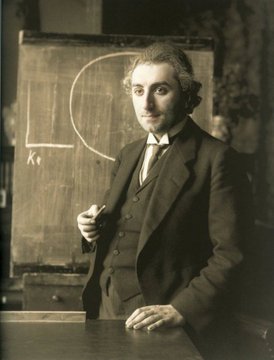 Sepia-toned historical photograph of a young man in a suit standing beside a chalkboard with geometric diagrams, holding chalk.