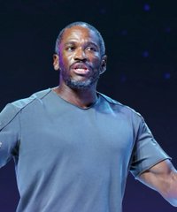A man with a short beard and shaved head, wearing a gray t-shirt, photographed from the chest up against a dark purple background.