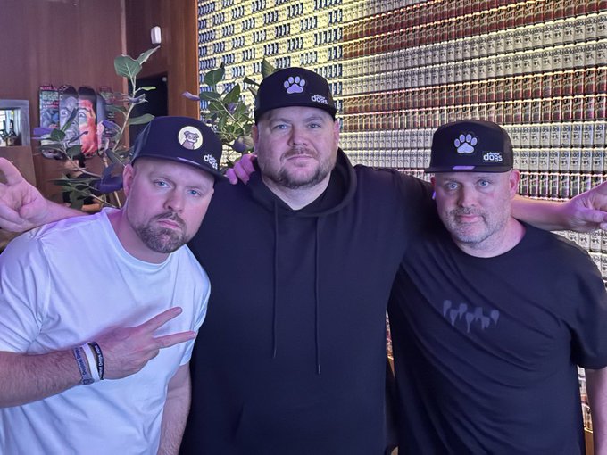 Three men pose together with arms around each other, all wearing black caps featuring a paw print logo and the text 'd&b'.