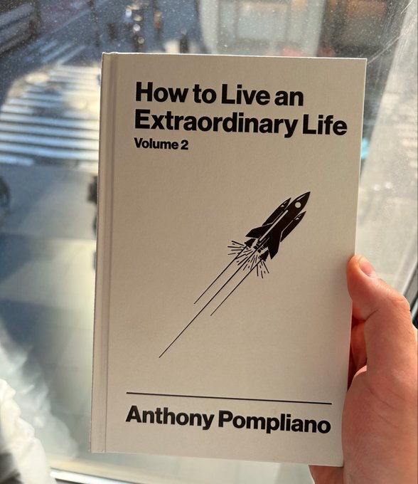A hand holding a book titled 'How to Live an Extraordinary Life Volume 2' by Anthony Pompiliano, featuring a rocket ship illustration on the cover.