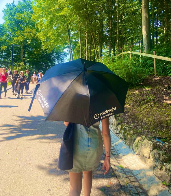 A person walking on a forest path holds a black 'midnight foundation' branded umbrella, with other hikers visible in the background.