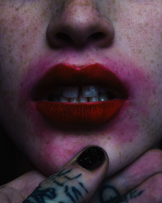 A close-up photograph of a person's lower face featuring red lipstick, freckled skin, and a hand with black nail polish and tattoos resting near the chin.