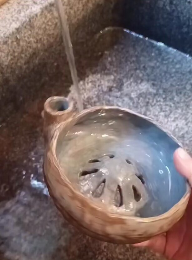 The image shows a person's hand holding a bowl of water, with some water splashing out from the faucet onto the countertop. The background appears to be a kitchen sink or counter area. There are no other objects visible in the image besides the bowl and the faucet.