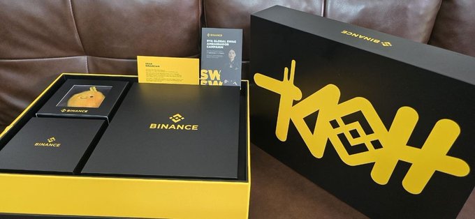 A black and yellow Binance 9th anniversary gift set sits on a brown leather couch, featuring a box with the Binance logo, a small Bitcoin coin, and a card honoring "CZ" for his role in building Binance's culture, alongside a large decorative yellow display piece.