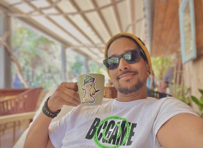 A man wearing a yellow beanie and sunglasses holding up a mug with a cartoon design, wearing a white 'BECAME' t-shirt.