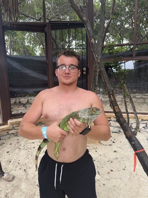 A shirtless man wearing glasses, black shorts, and a wristband stands on a sandy beach holding a large green iguana, with a fenced enclosure and trees in the background.