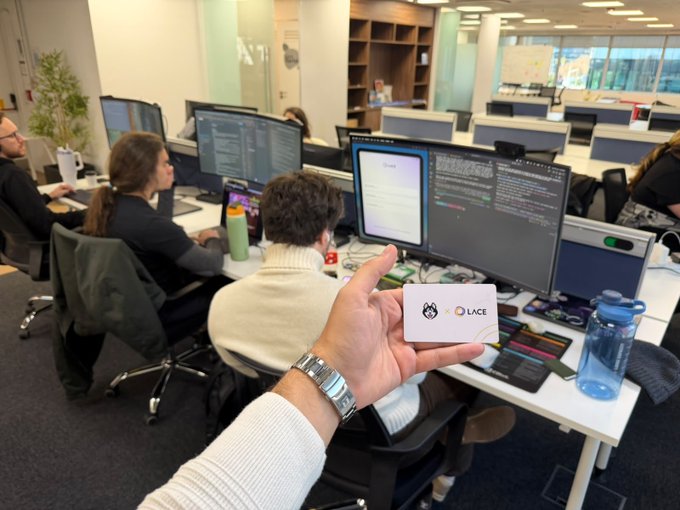 The image shows a group of people working together in an office setting with multiple computers and monitors. One person is holding up a white business card that says "lace" on it, possibly indicating their involvement or affiliation with the company. The office appears to be a shared workspace where individuals collaborate and work on various projects.
