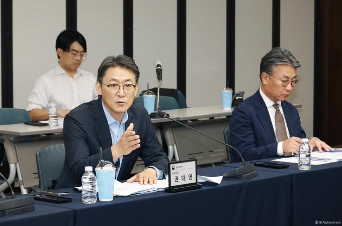 A man in a dark suit speaks at a conference table with a nameplate reading 'Kwon Deok-yeong' in Korean, while two other men in suits sit nearby.