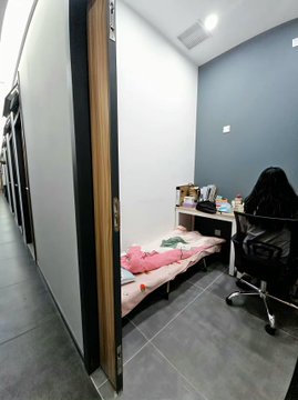 A compact, minimalist workspace featuring a small desk with a laptop, a chair, and a simple bed with pink bedding tucked into a narrow corner room, reflecting the "1 person, 1 computer, 1 desk, 1 phone" ultra-lean startup model of Shenzhen OPCs.