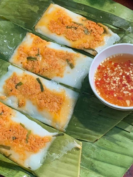 Rectangular pieces of white steamed rice cake topped with orange crumbs are served on banana leaves, accompanied by a small bowl of red chili dipping sauce with sesame seeds.