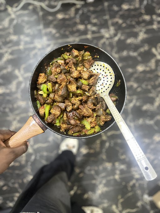 A hand holds a wooden-handled pan filled with a cooked meat and green pepper dish, with a slotted spoon resting inside.